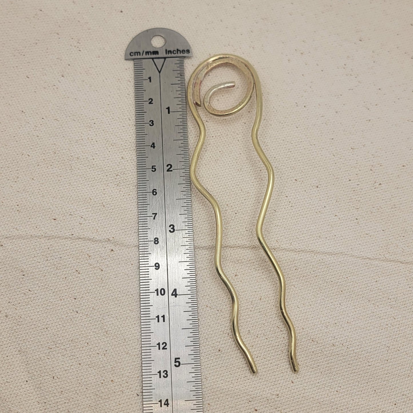 Brass hair pin next to a ruler on a textured beige surface
