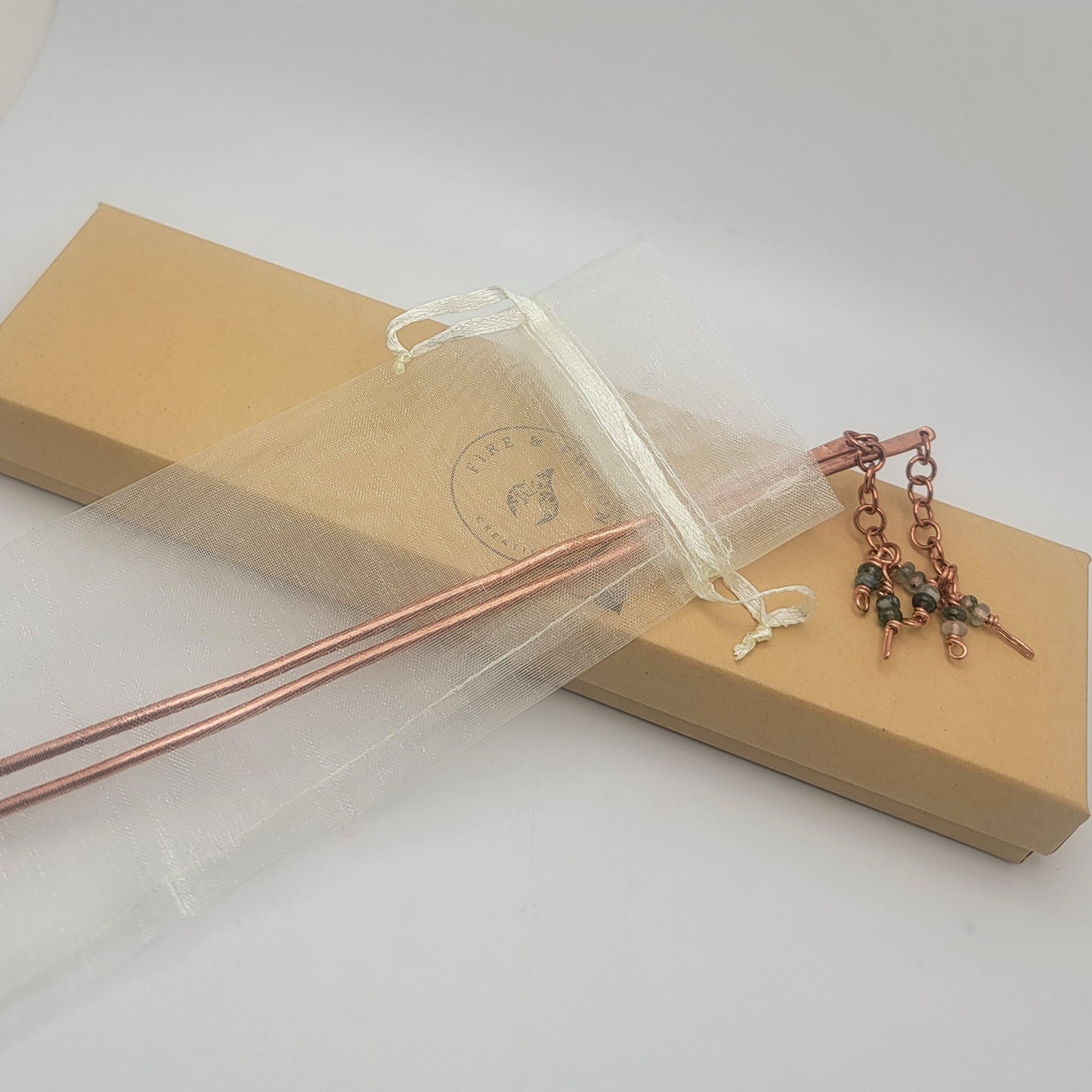 A pair of copper beaded hair sticks in an organza gift bag on a long cardboard gift box on a white background