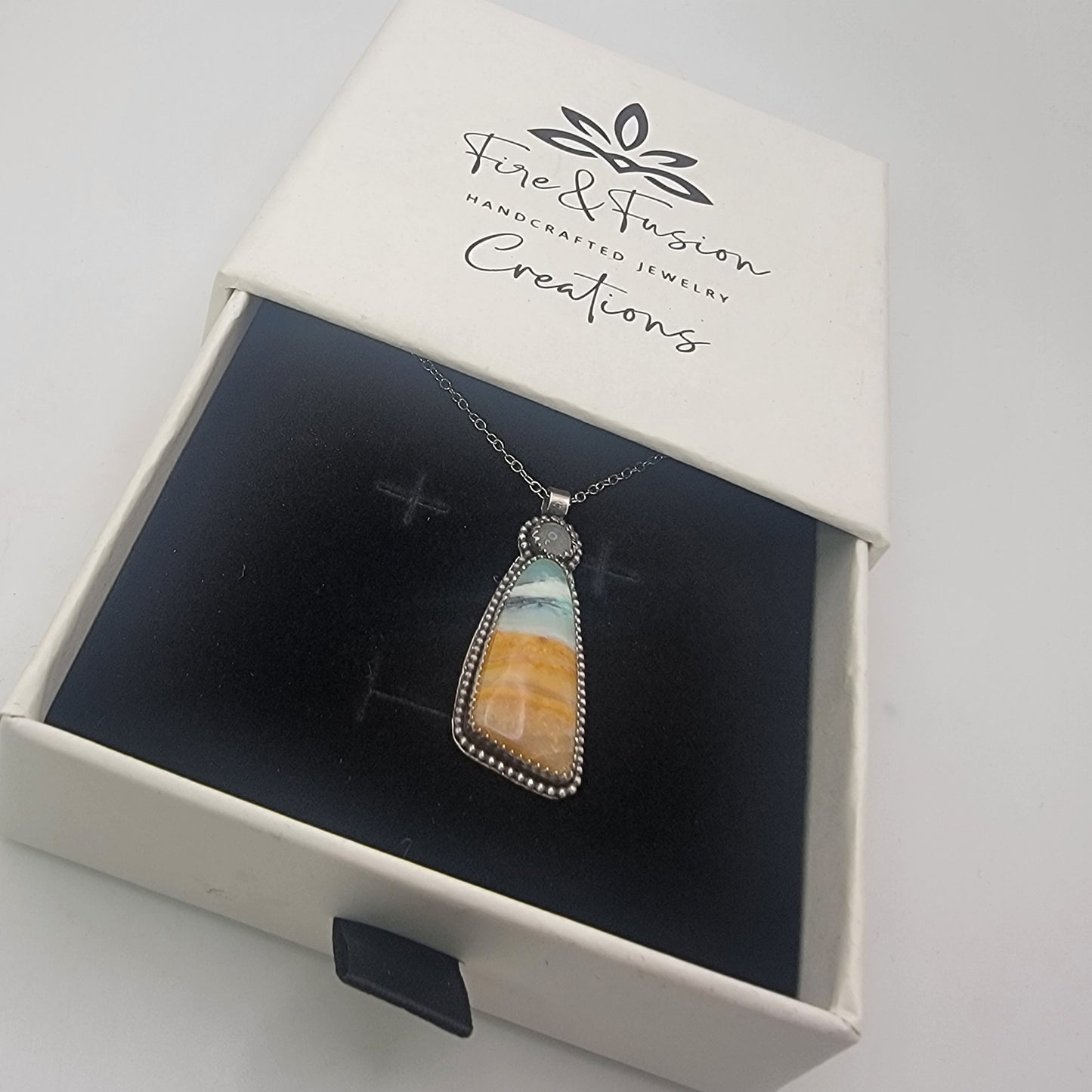 A silver-toned pendant necklace with a triangular stone showing blue, white, and orange banding, displayed inside an open white jewelry box branded 'Fire & Fusion Handcrafted Jewelry Creations'.