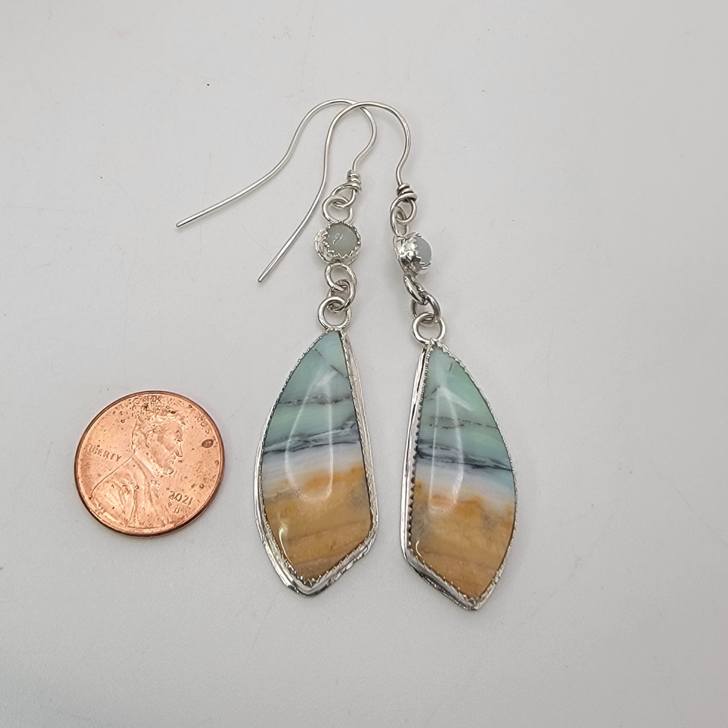 A pair of dangling earrings with irregular marquise-shaped resin pendants showing a marbled pattern of teal, gray, and ochre orange, suspended from silver fishhook wires. A US quarter coin is placed to the left for scale, set against a plain light gray background.