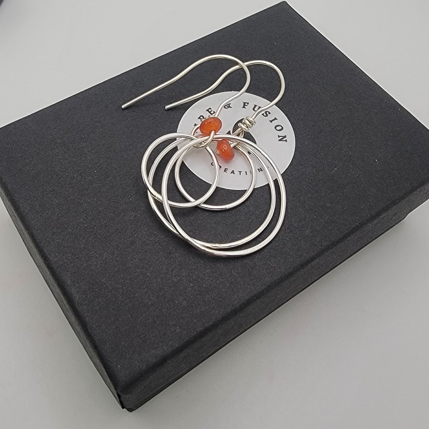 A pair of silver-toned drop earrings with small orange-red bead accents, presented on a white circular card atop a black square box against a light gray background.