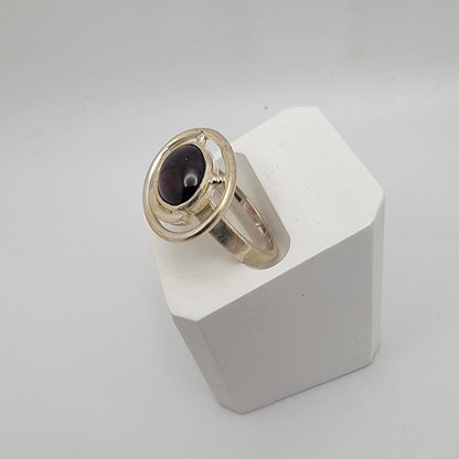 Sterling silver ring with a 12×10 mm oval garnet cabochon set on a uniform 3 mm band, displayed on a white stand.