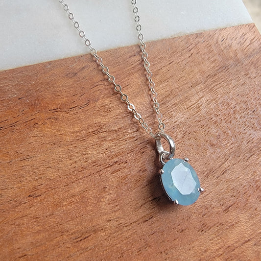 Faceted oval aquamarine gemstone pendant in sterling silver four-prong setting on silver chain, displayed on wood and marble background.