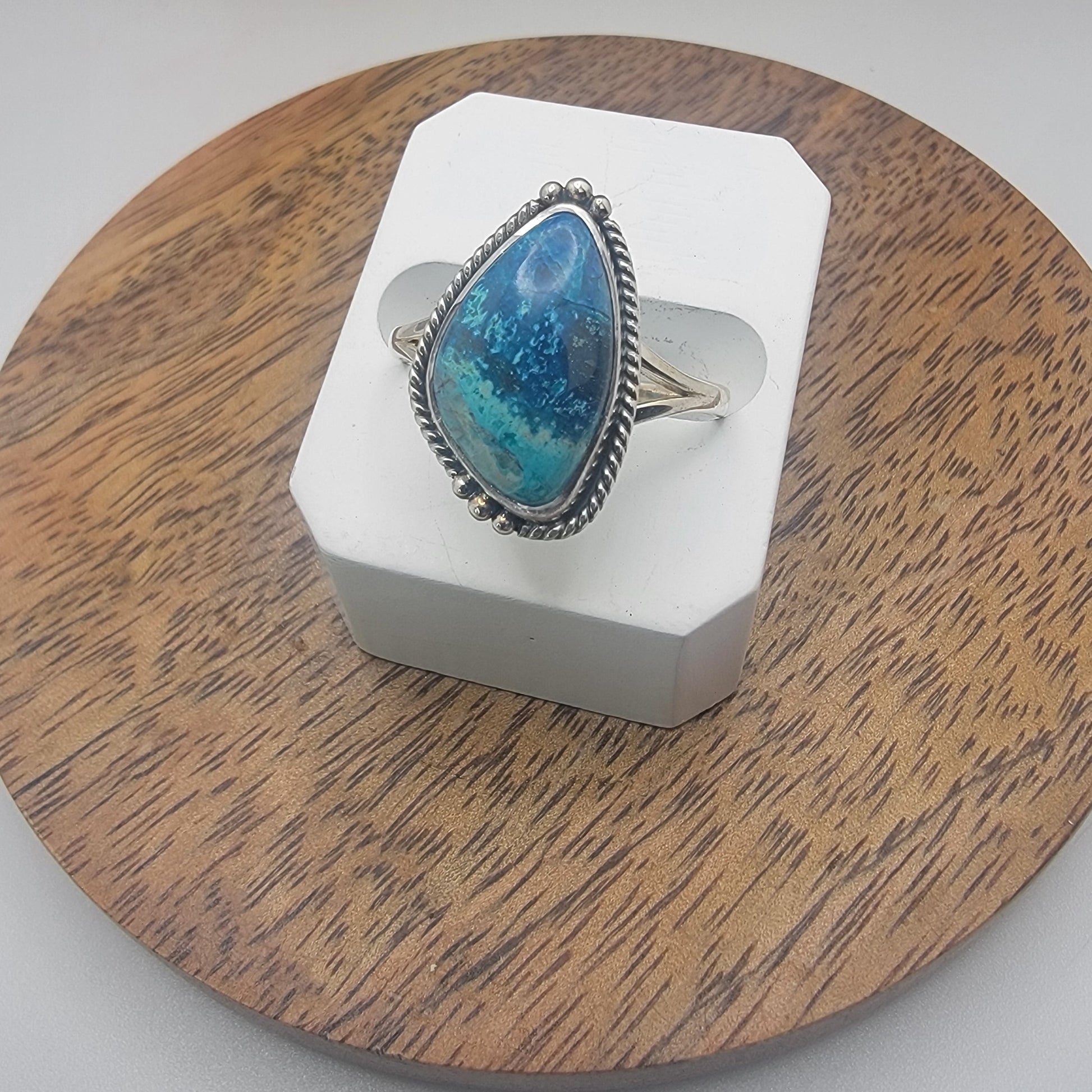 Freeform shaped shattuckite gemstone ring in sterling silver with rope edge and bead detail, featuring vibrant blue and turquoise hues, displayed on white stand.