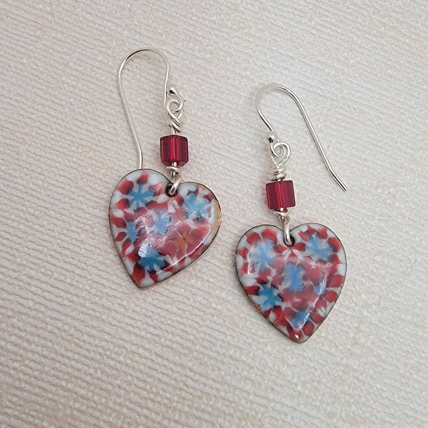 Heart-shaped earrings with floral pattern and red beads on a textured beige surface