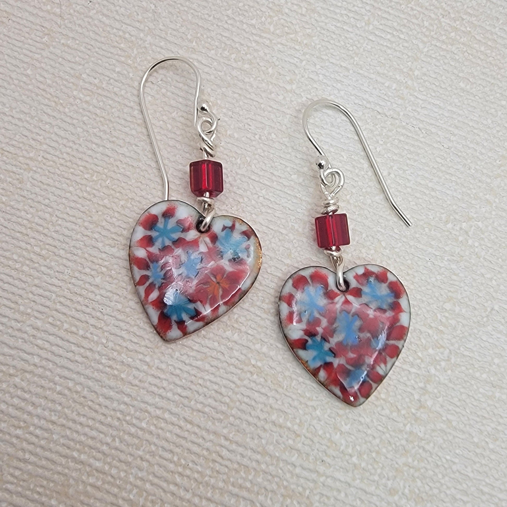 Heart-shaped earrings with floral pattern and red beads on a textured beige surface