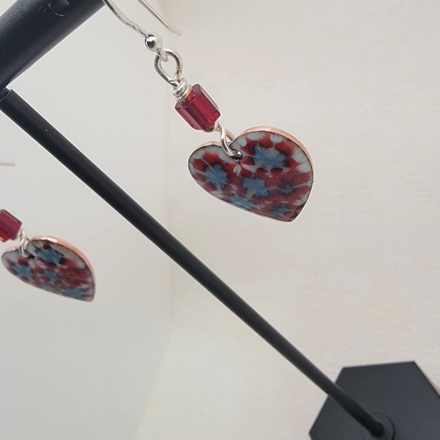Heart-shaped earrings with red and blue patterns on a black stand.