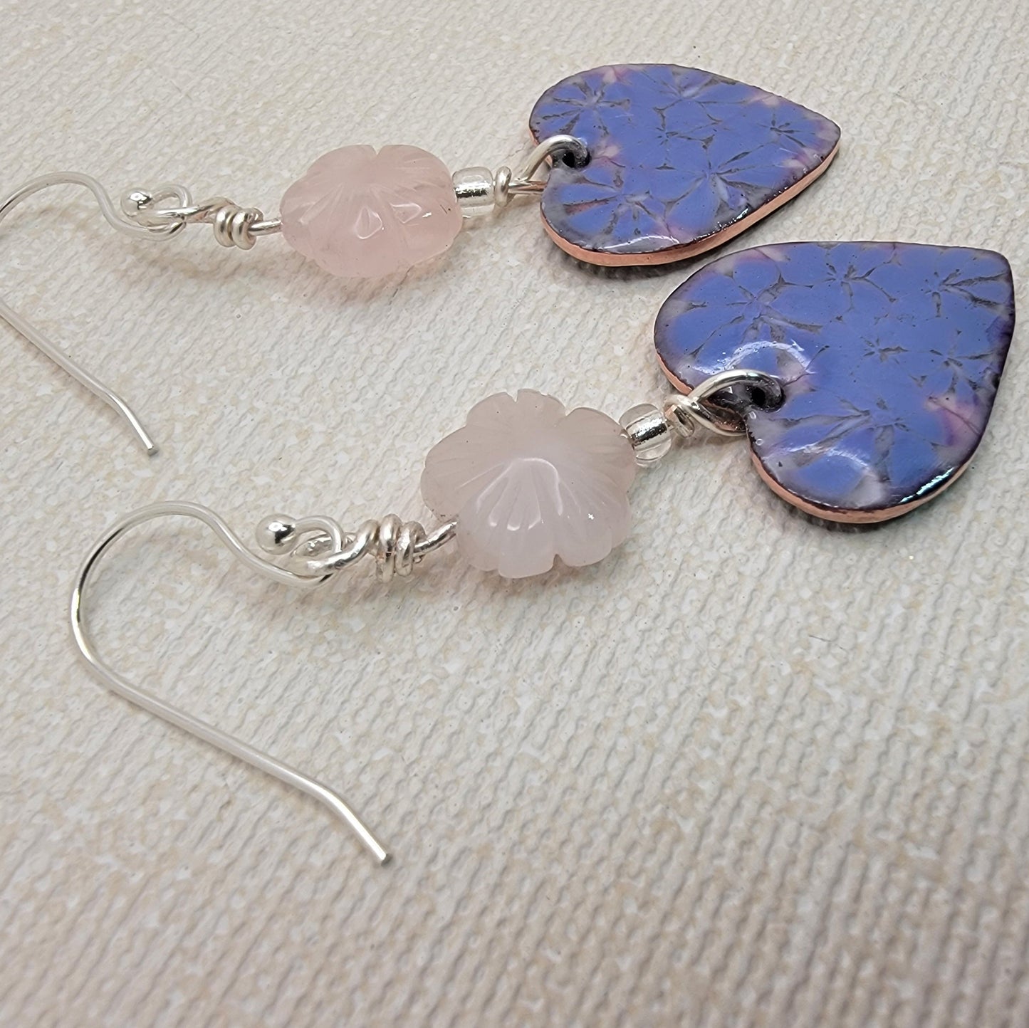 Earrings with purple heart-shaped stones and pink rose quartz beads on a textured beige surface.
