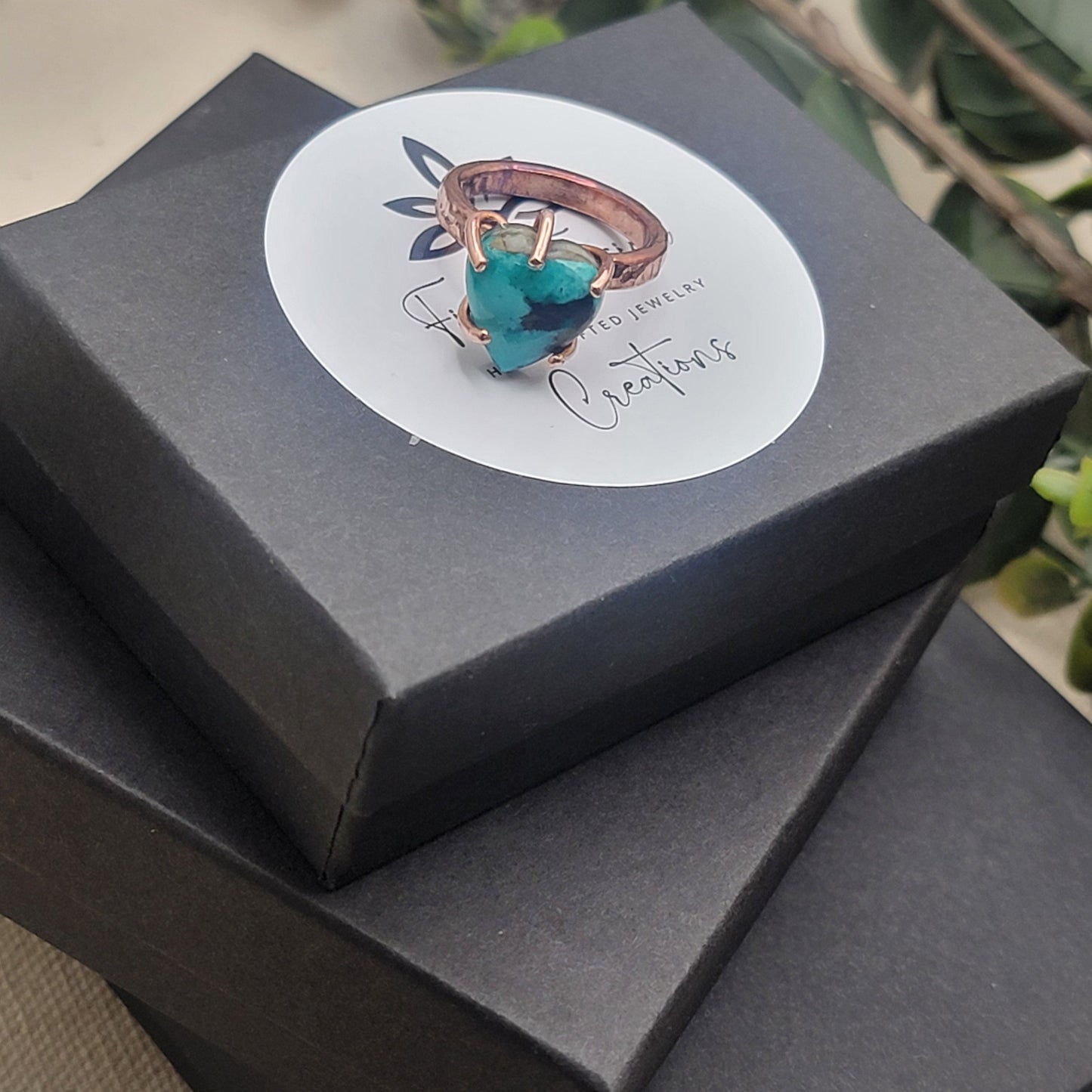 Copper and turquoise ring on gift box stack next to plant on beige background