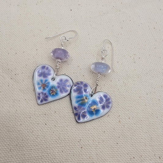 White, purple and blue enameled heart earrings with purple lampworked glass bead and silver ear wires on a beige background