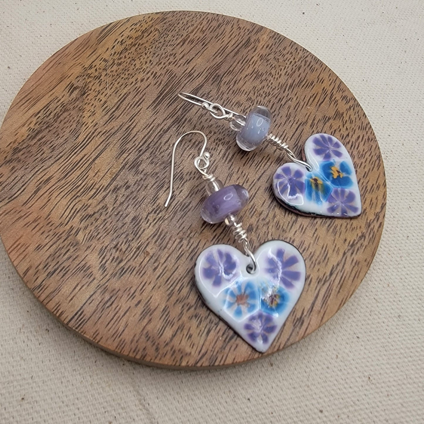White, purple and blue enameled heart earrings with purple lampworked glass bead and silver ear wires on a round wood platform on a beige background