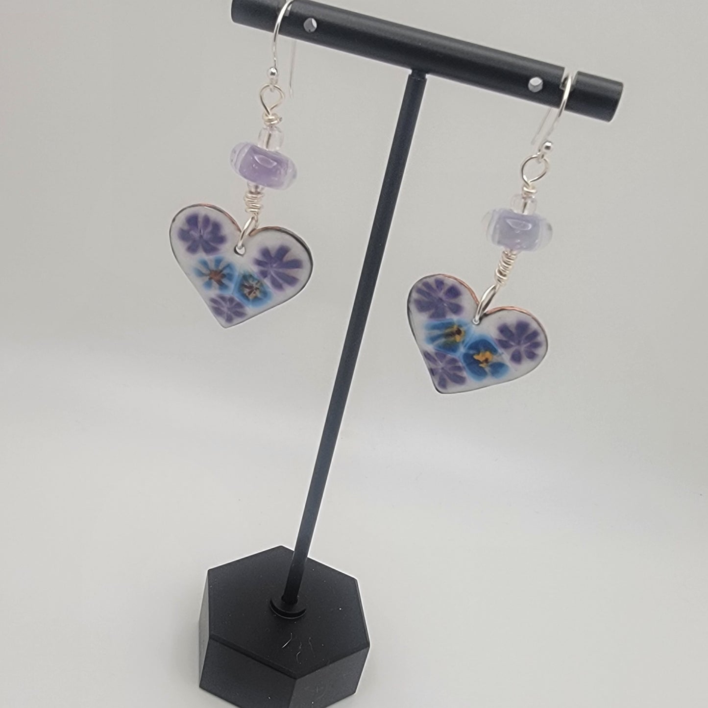 White, purple and blue enameled heart earrings with purple lampworked glass bead and silver ear wires dangling from a black earring display on a beige background