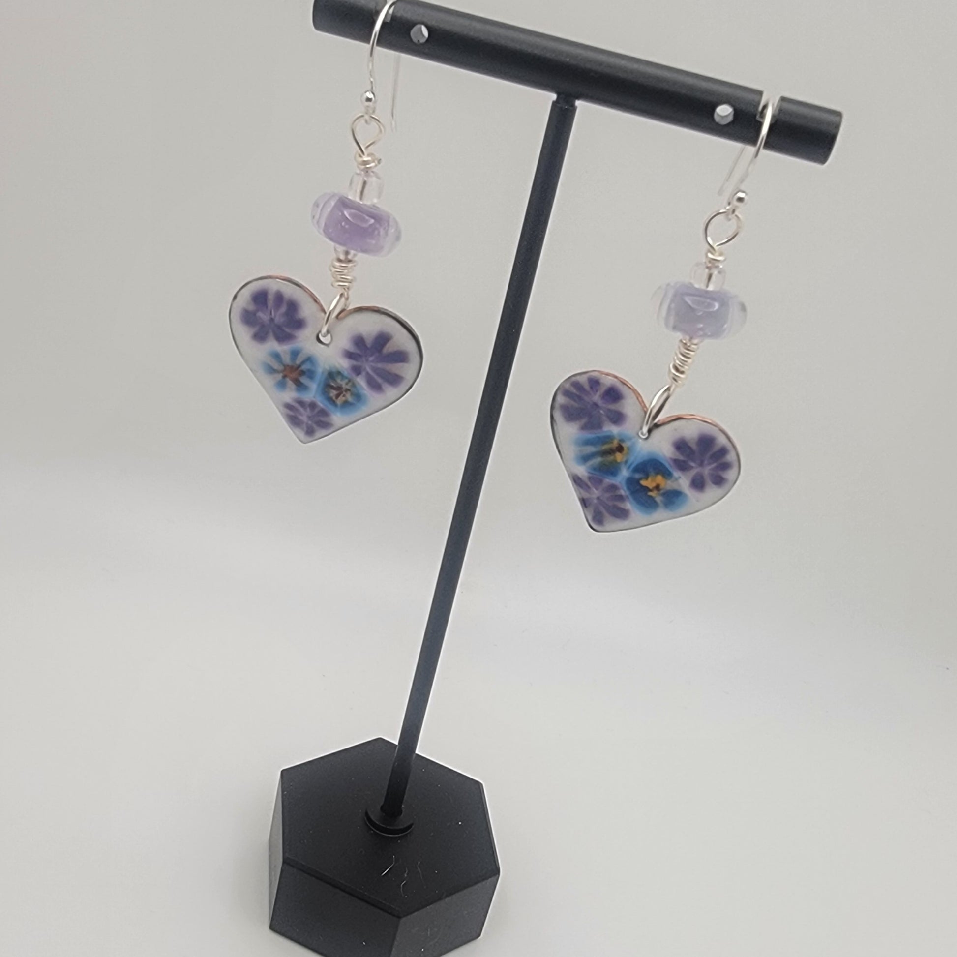 White, purple and blue enameled heart earrings with purple lampworked glass bead and silver ear wires dangling from a black earring display on a beige background