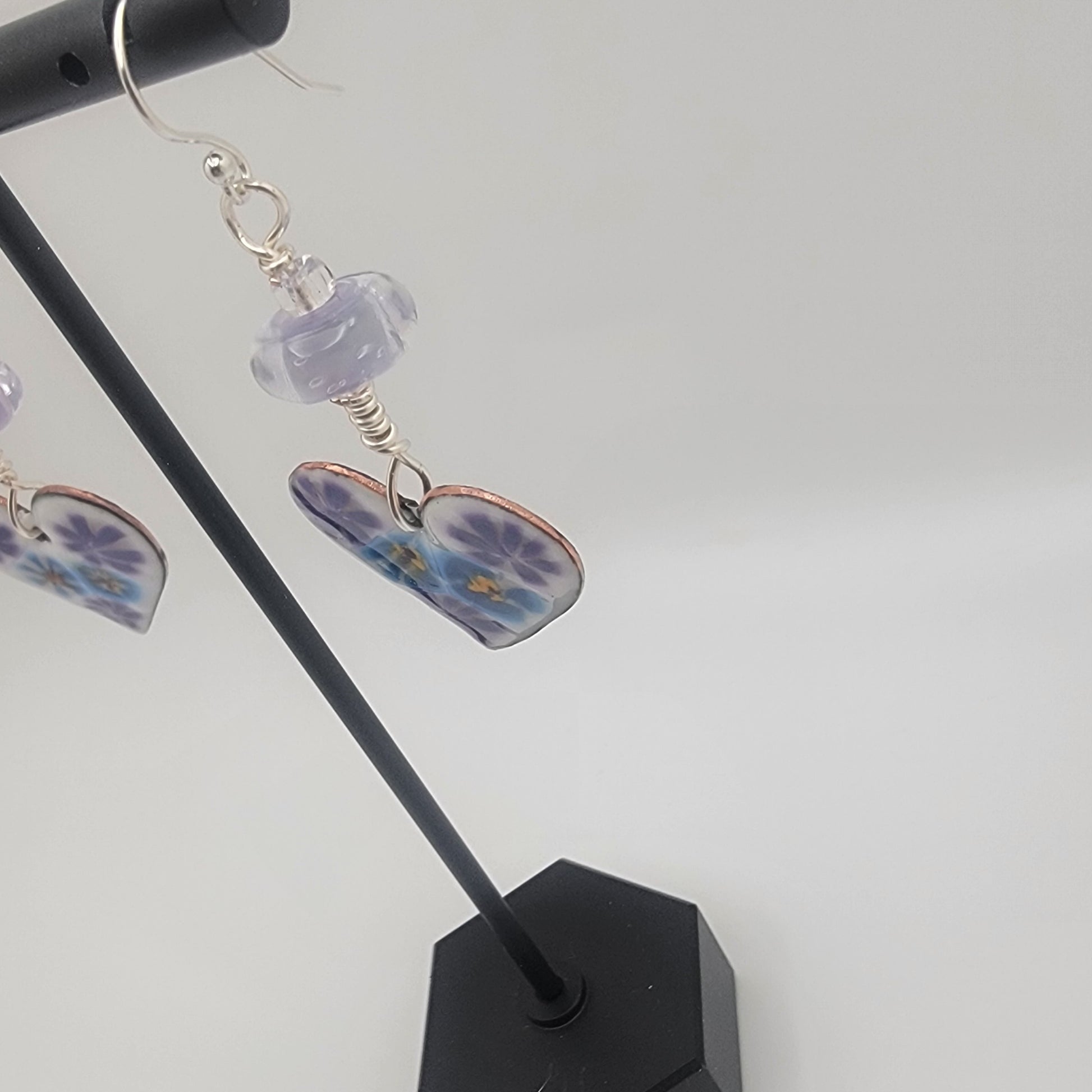 White, purple and blue enameled heart earrings with purple lampworked glass bead and silver ear wires dangling from a black earring display on a beige background