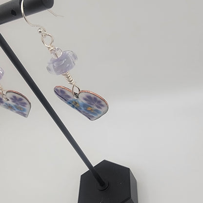 White, purple and blue enameled heart earrings with purple lampworked glass bead and silver ear wires dangling from a black earring display on a beige background