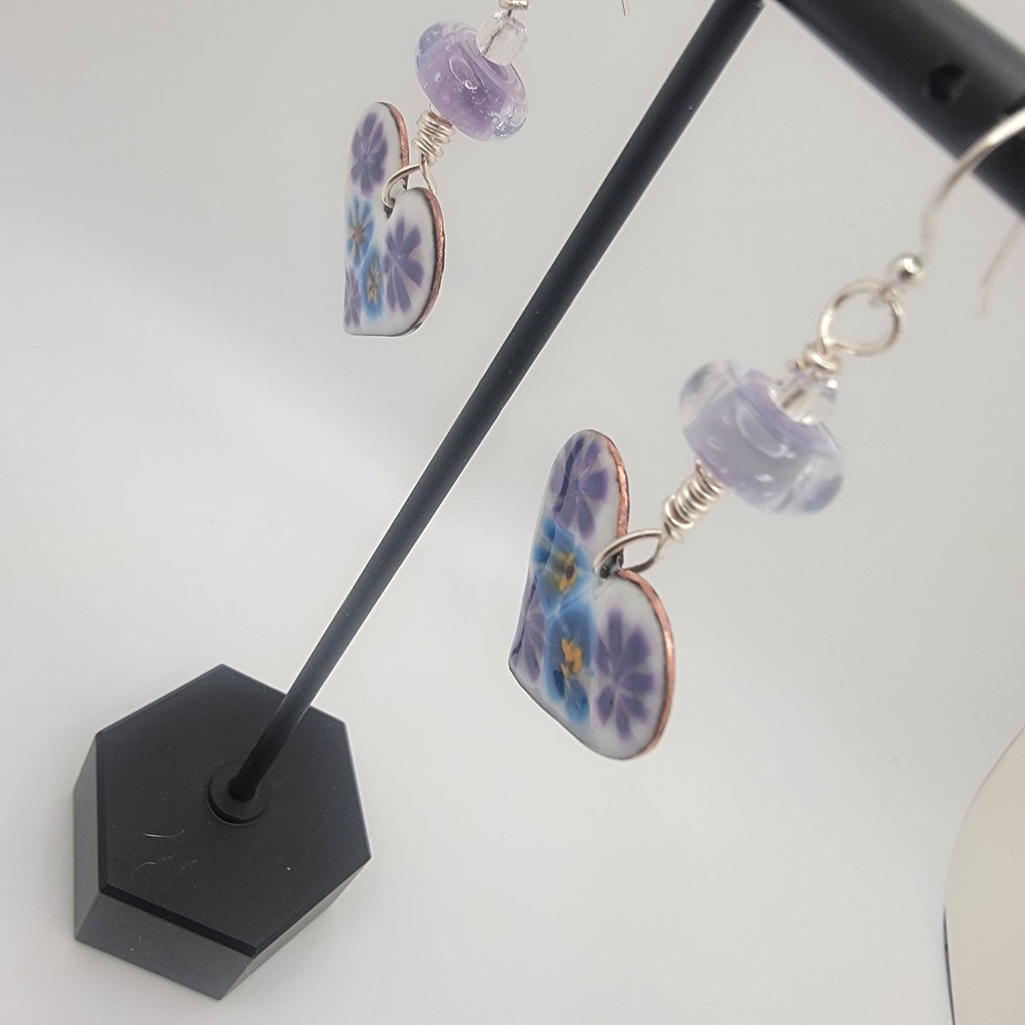 White, purple and blue enameled heart earrings with purple lampworked glass bead and silver ear wires dangling from a black earring display on a beige background