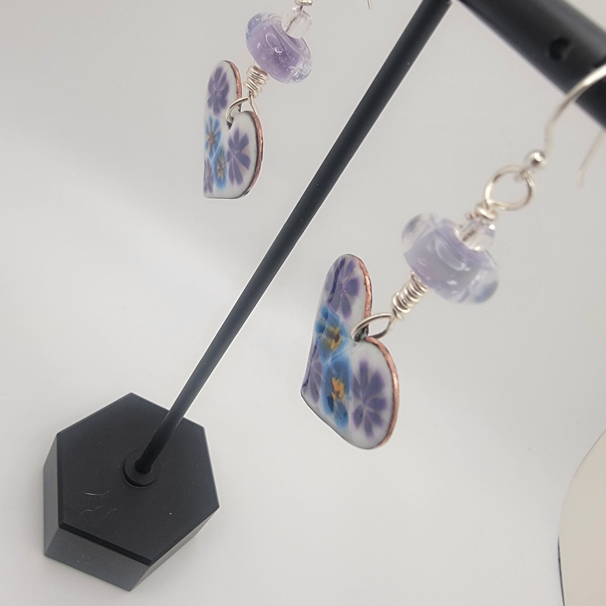 White, purple and blue enameled heart earrings with purple lampworked glass bead and silver ear wires dangling from a black earring display on a beige background
