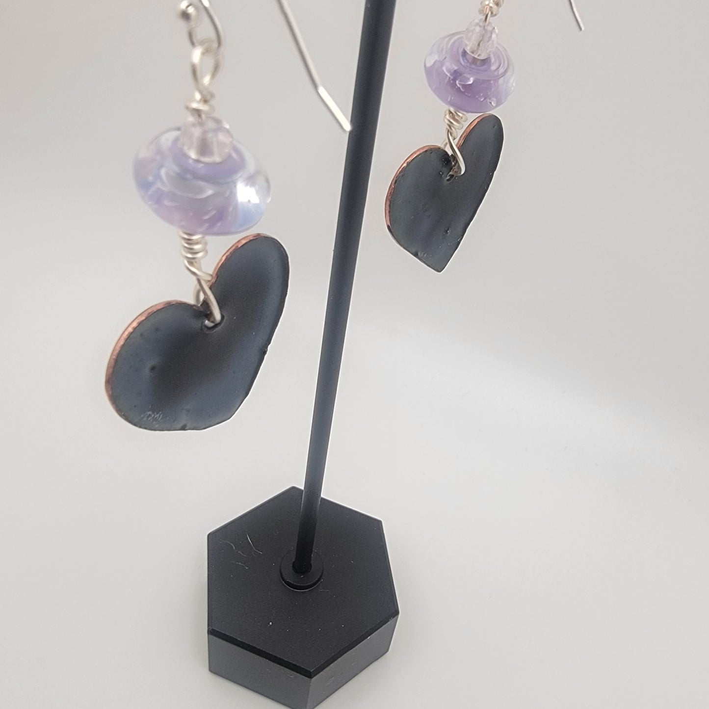 White, purple and blue enameled heart earrings with purple lampworked glass bead and silver ear wires dangling from a black earring display on a beige background