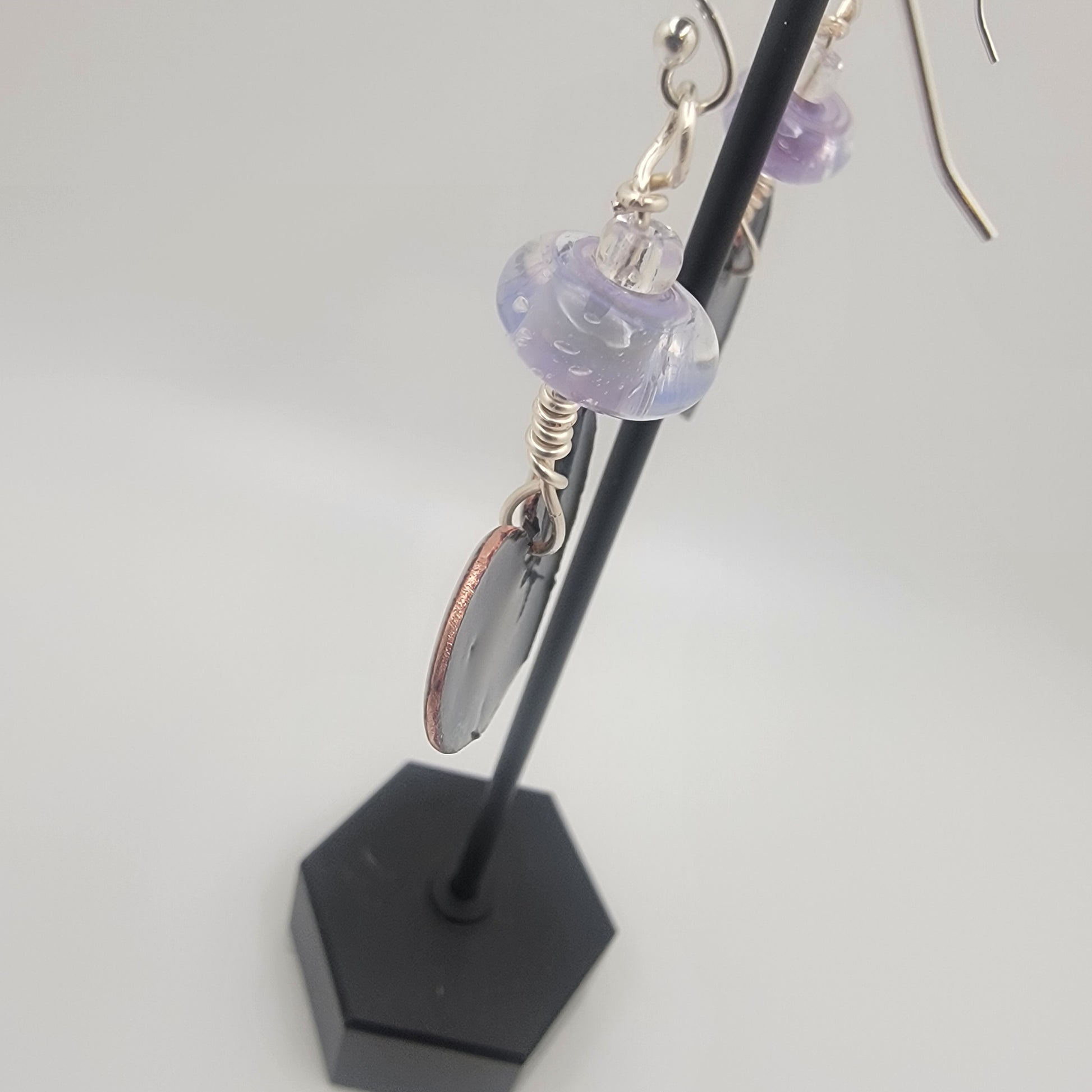 White, purple and blue enameled heart earrings with purple lampworked glass bead and silver ear wires dangling from a black earring display on a beige background