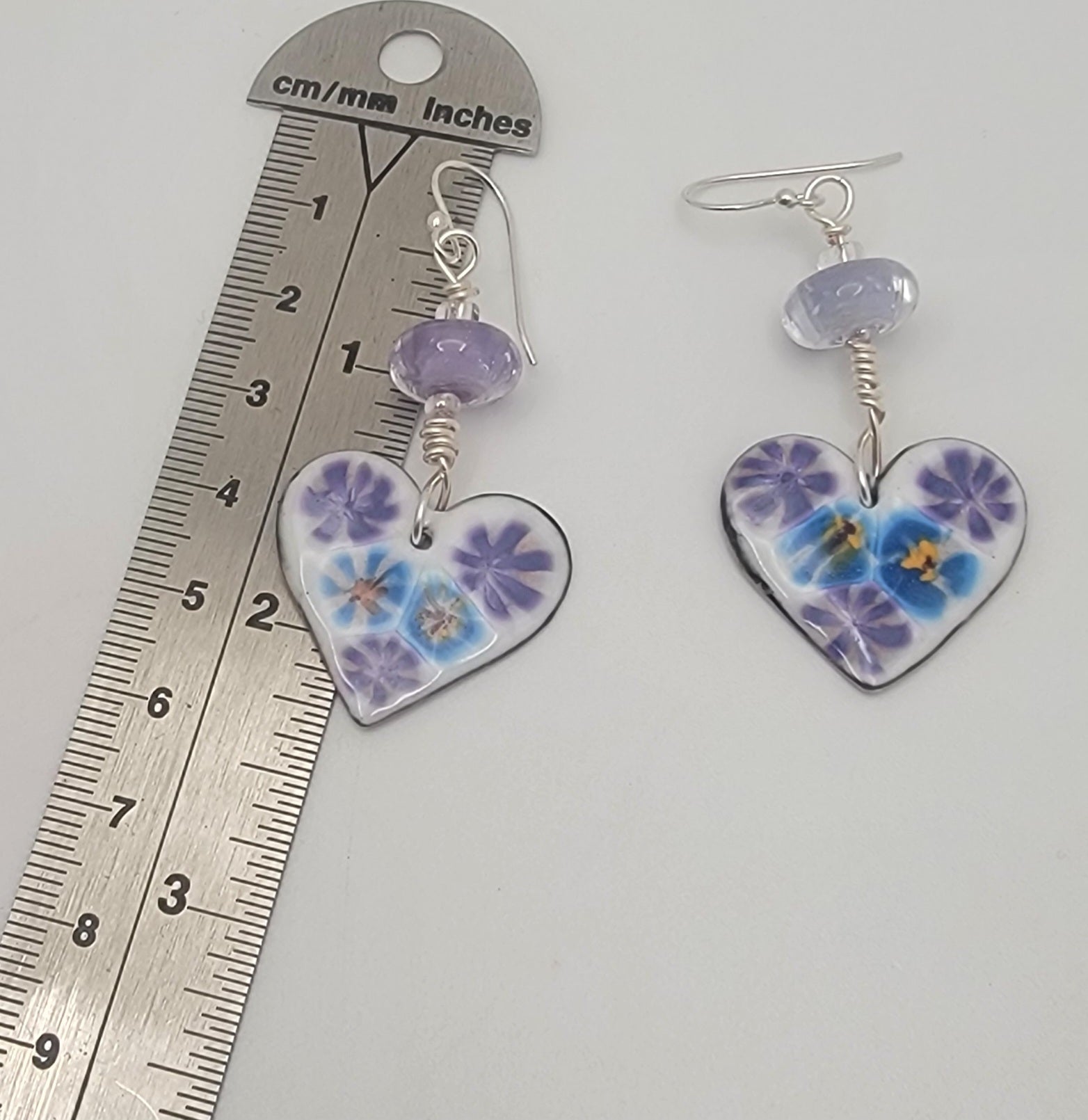 White, purple and blue enameled heart earrings with purple lampworked glass bead and silver ear wires on a beige background next to a ruler