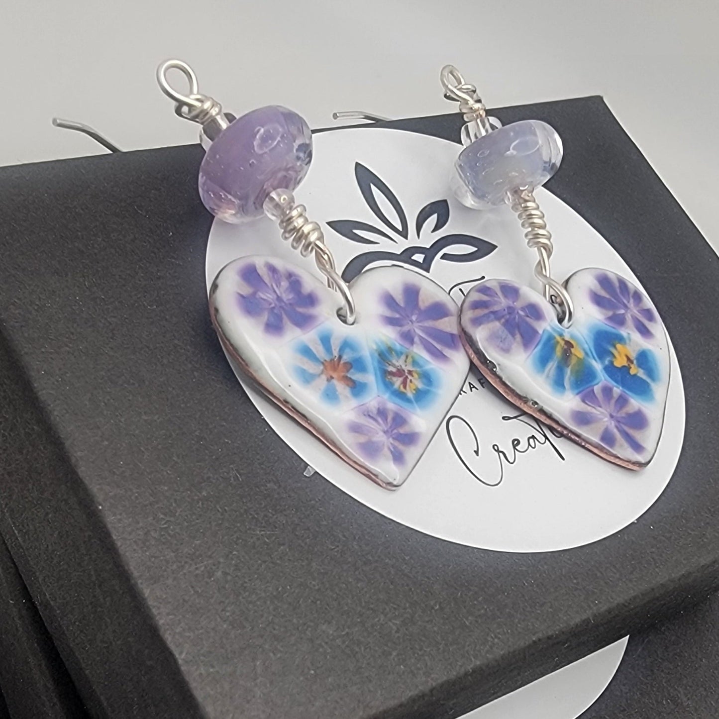 White, purple and blue enameled heart earrings with purple lampworked glass bead and silver ear wires on a stack of black gift boxes on a beige background