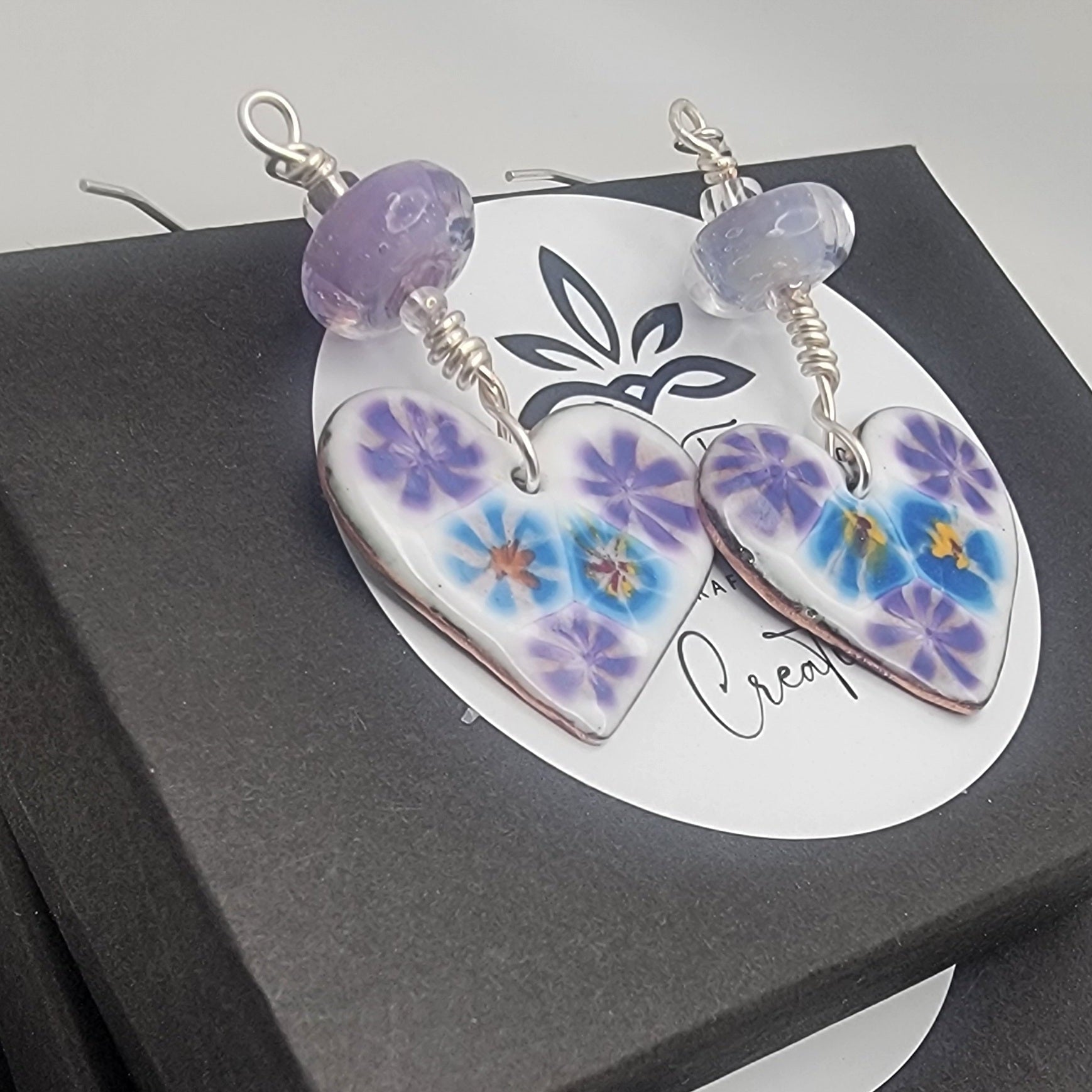 White, purple and blue enameled heart earrings with purple lampworked glass bead and silver ear wires on a stack of black gift boxes on a beige background