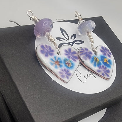 White, purple and blue enameled heart earrings with purple lampworked glass bead and silver ear wires on a stack of black gift boxes on a beige background