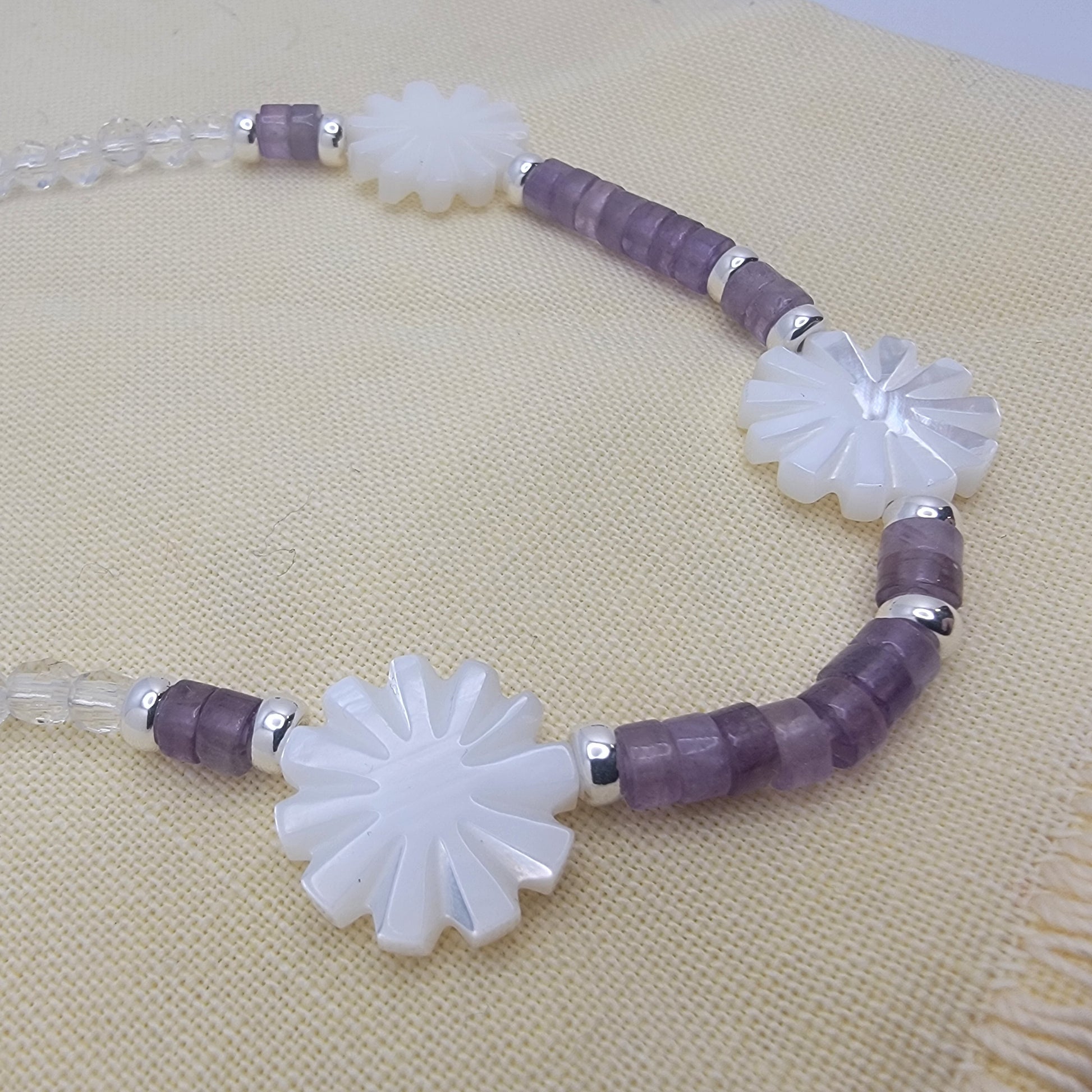 Necklace with white floral beads and purple and clear cylindrical beads on a beige fabric background