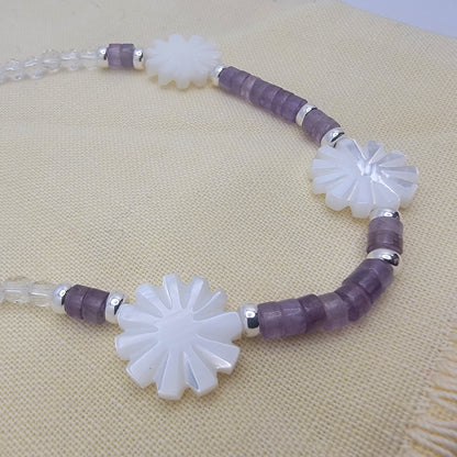 Necklace with white floral beads and purple and clear cylindrical beads on a beige fabric background