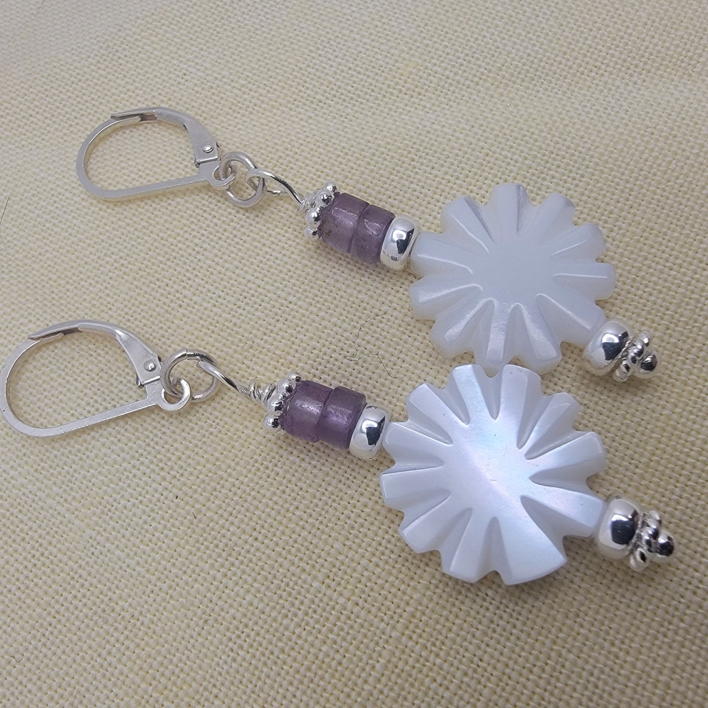 Pair of earrings with white floral designs and purple beads on a beige fabric background