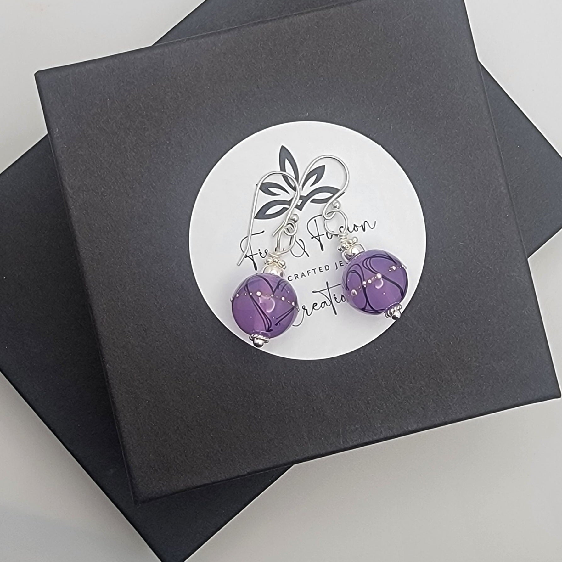 Purple earrings on a card with a brand logo, placed on black boxes.