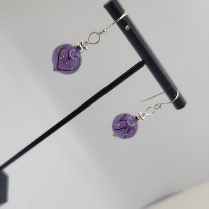 Purple heart-shaped earrings on a stand against a light gray background