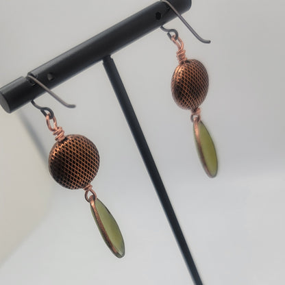 Pair of earrings with copper mesh and green teardrop shapes on a stand.