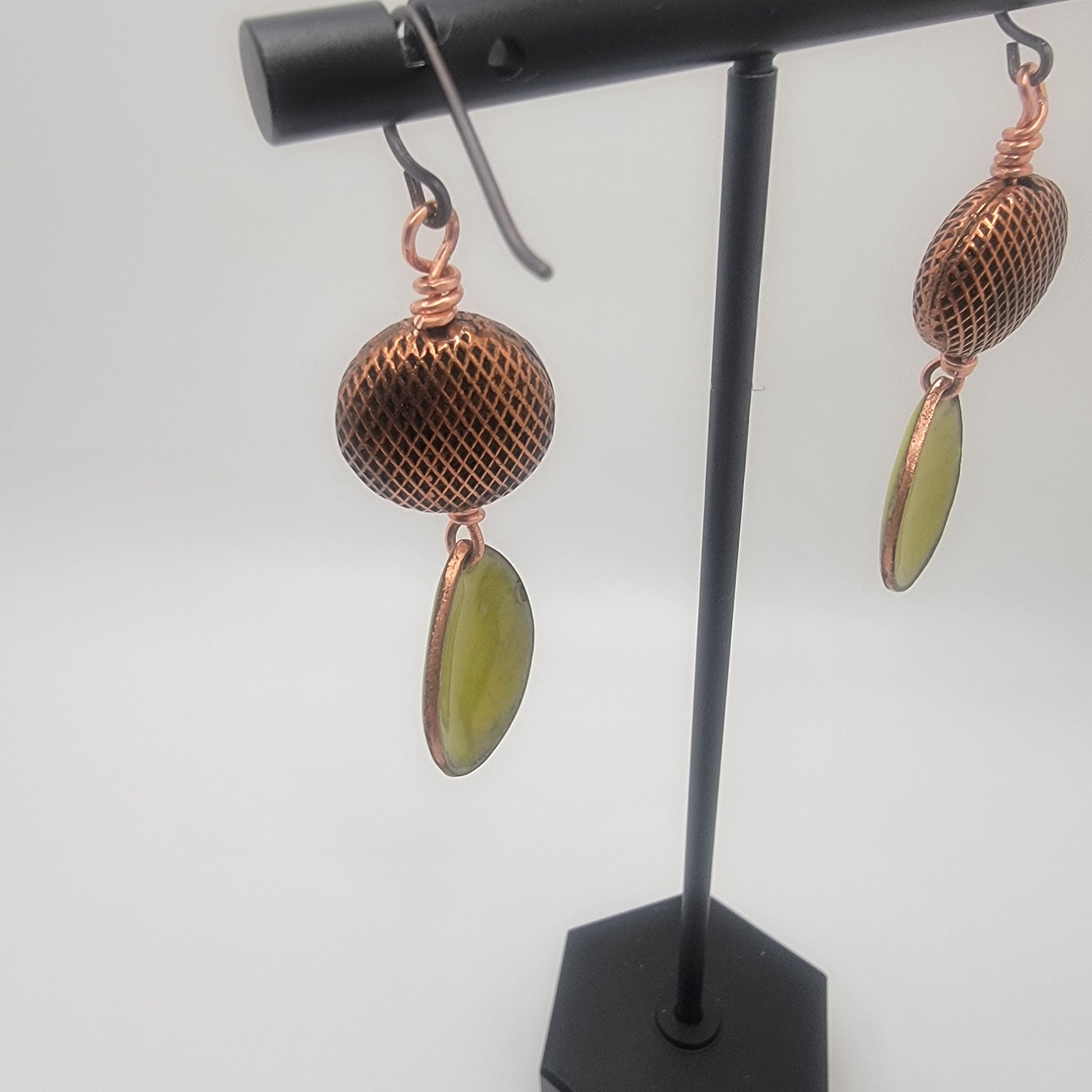 Pair of earrings with copper-like finish and greenish leaves on a black stand against a white background