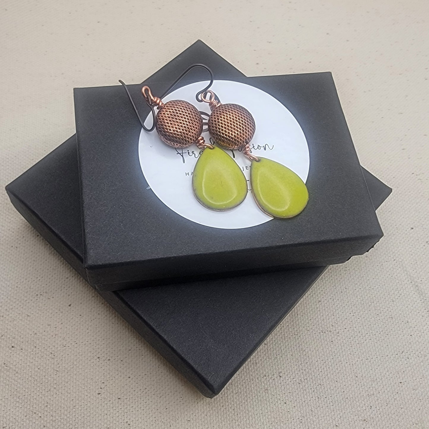 Pair of earrings with green teardrop-shaped stones and bronze circular tops on a black box.