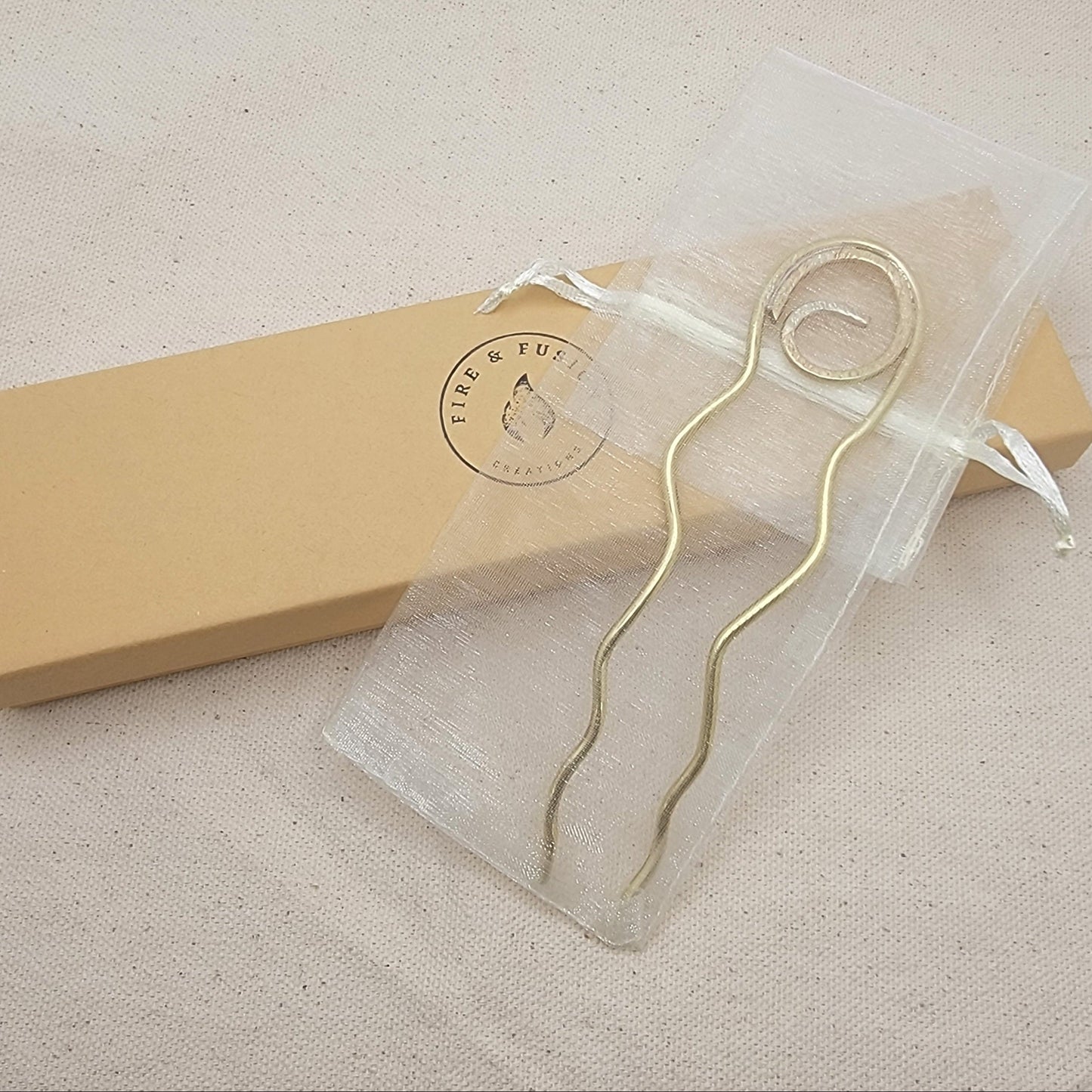 Brass hair pin in organza bag next to branded gift box with a beige background