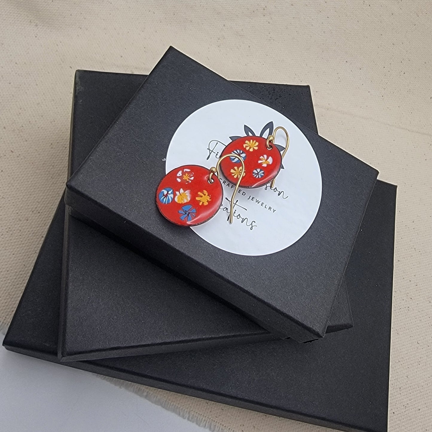 Black jewelry box with a round label featuring red floral earrings on a beige surface.