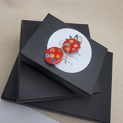 Black jewelry box with a round label featuring red floral earrings on a beige surface.