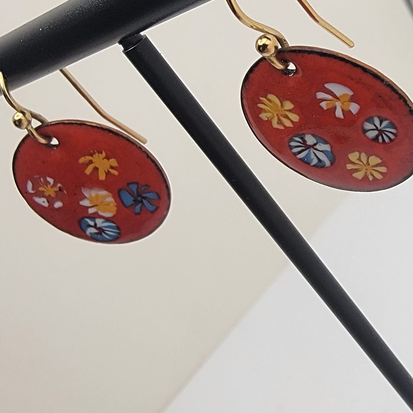 Pair of earrings with red circular pendants featuring colorful floral designs on a black stand.