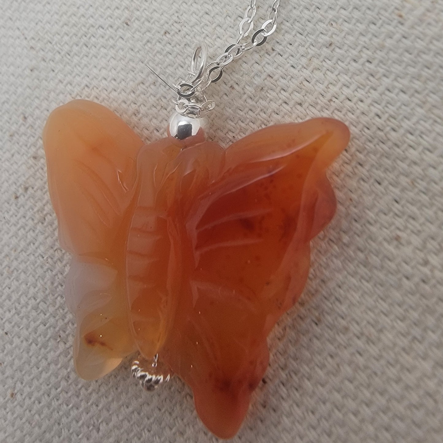 Butterfly-shaped orange pendant on a chain against a textured beige background
