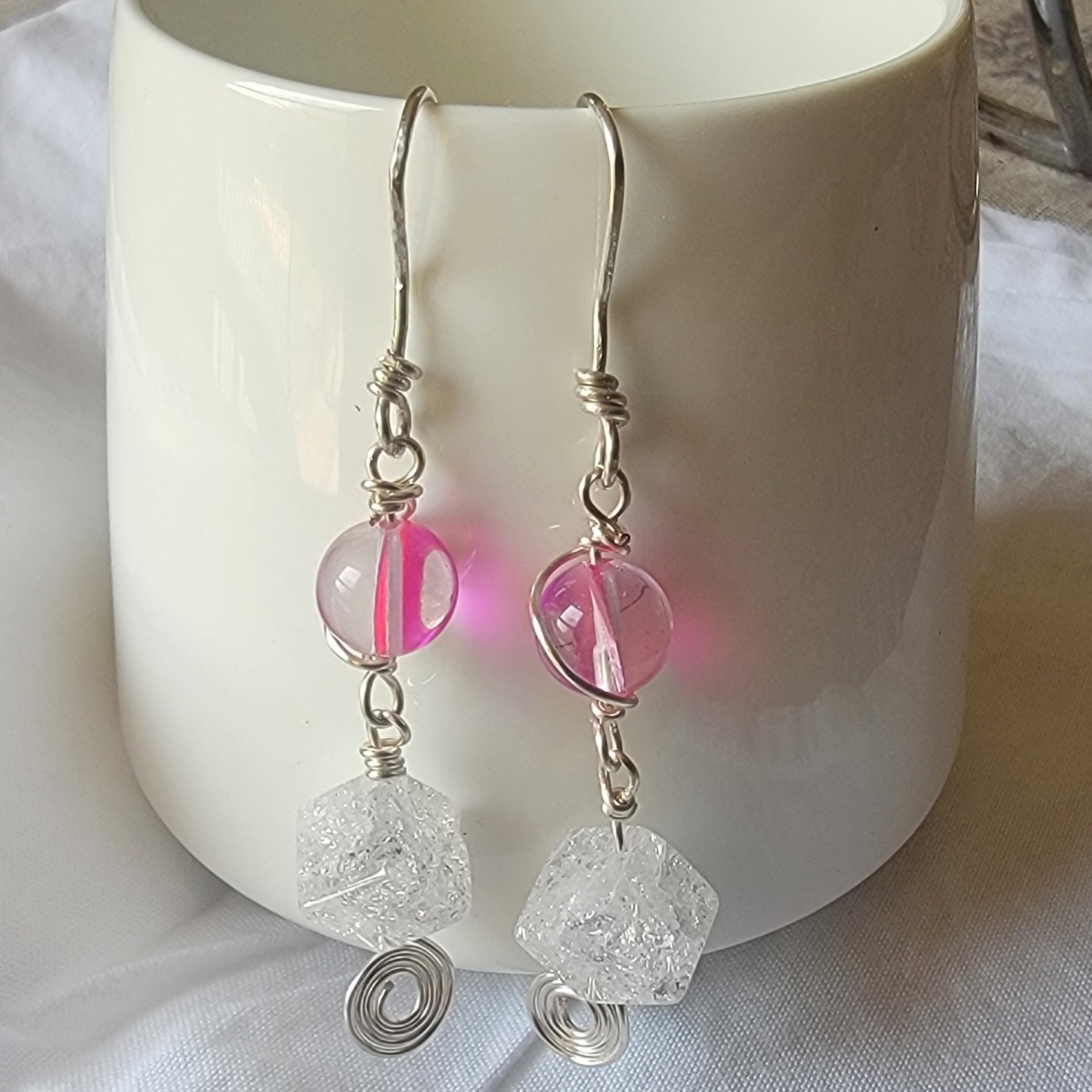 Silver earrings with pink and clear beads hanging in front of a white mug.