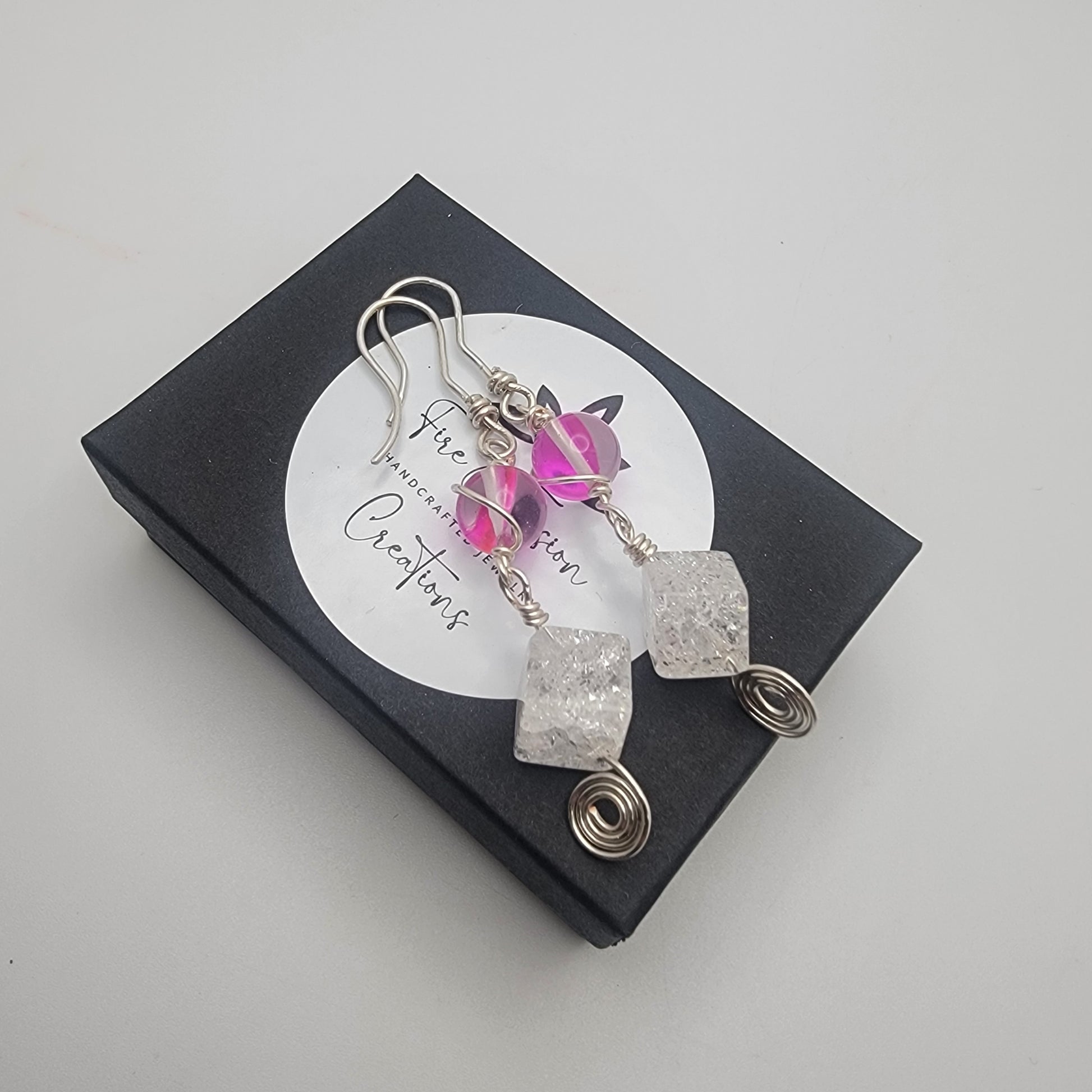 Earrings with pink gemstone and silver wire on a black box with 'The Gemstone Creations' label.