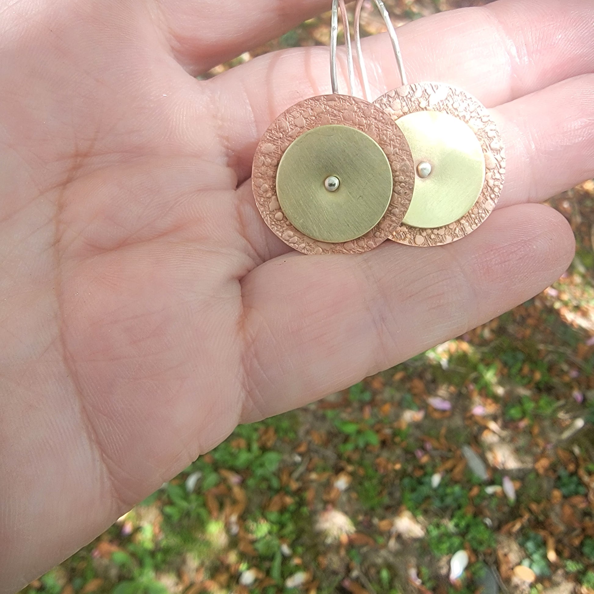 Copper and brass circular earrings held in a hand with a blurred natural background
