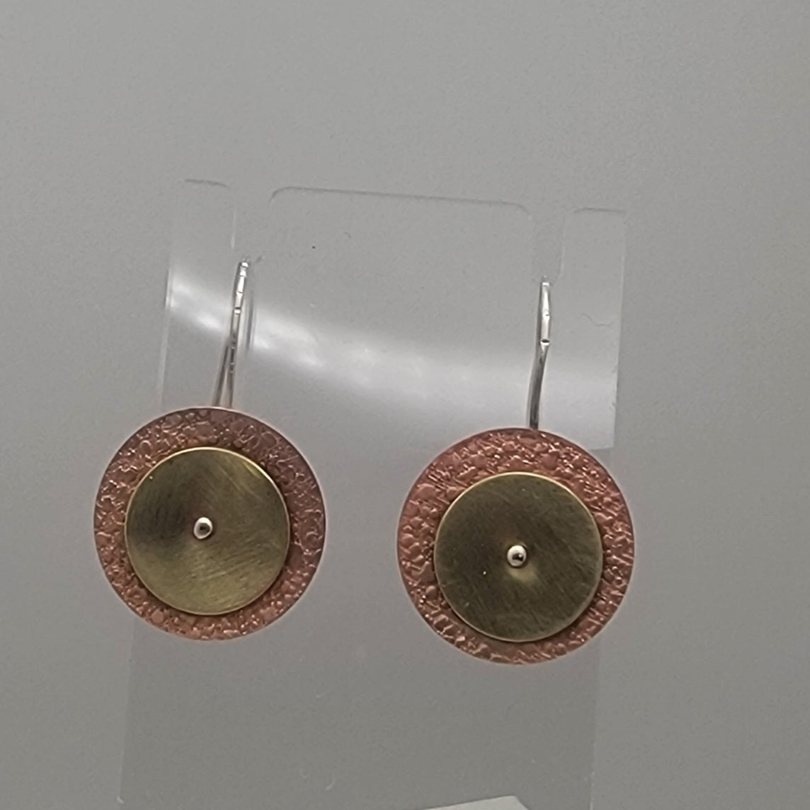 Pair of gold and bronze circular earrings on a clear stand against a gray background