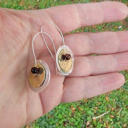 Silver and brass earrings held in a hand with a green outdoor background