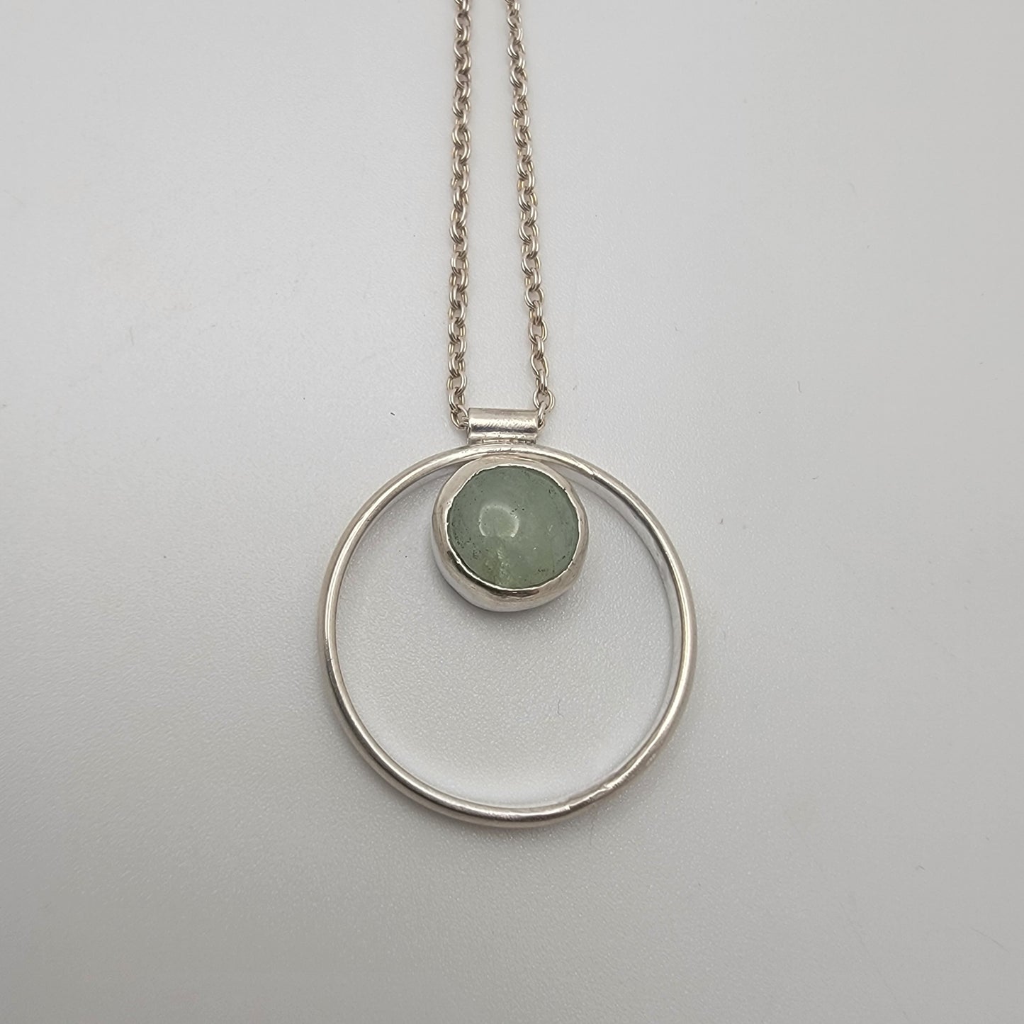 Silver necklace with a circular pendant featuring a green gemstone on a light gray background