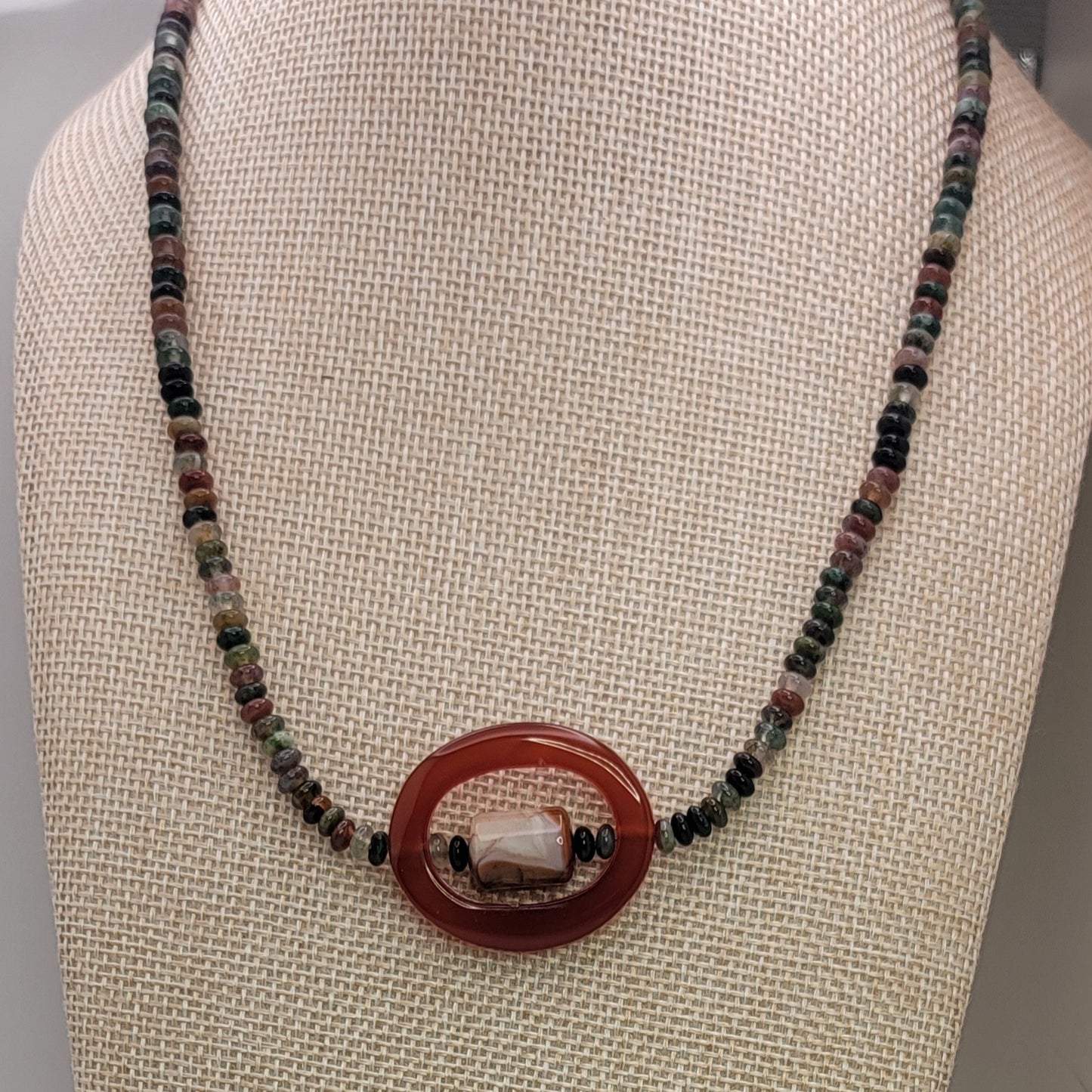 Beaded necklace with small earth tone beads and large oval carnelian bead on a beige necklace display