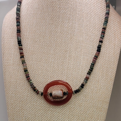 Beaded necklace with small earth tone beads and large oval carnelian bead on a beige necklace display