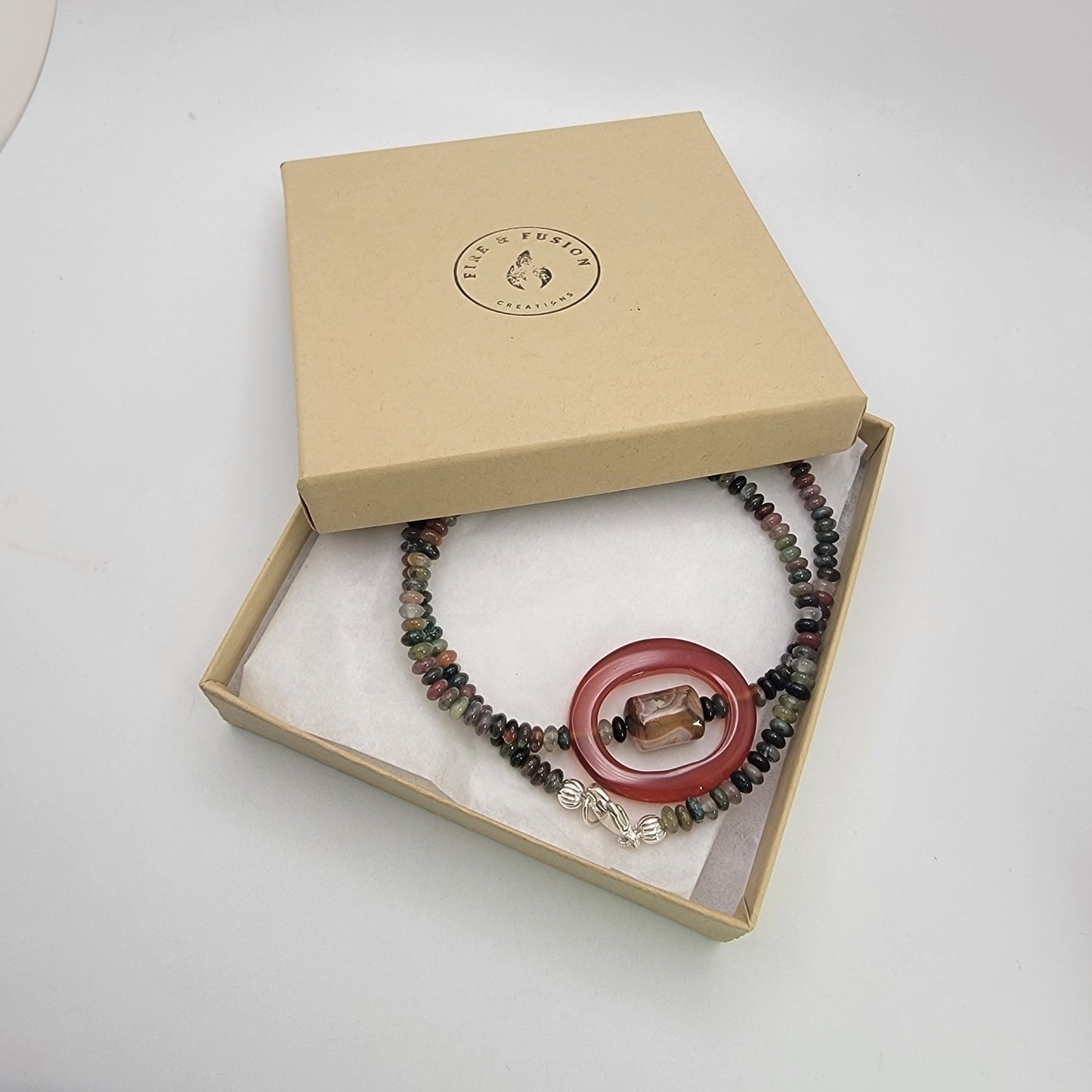 Beaded necklace with small earth tone beads and large oval carnelian bead in a neutral color branded gift box and a white background