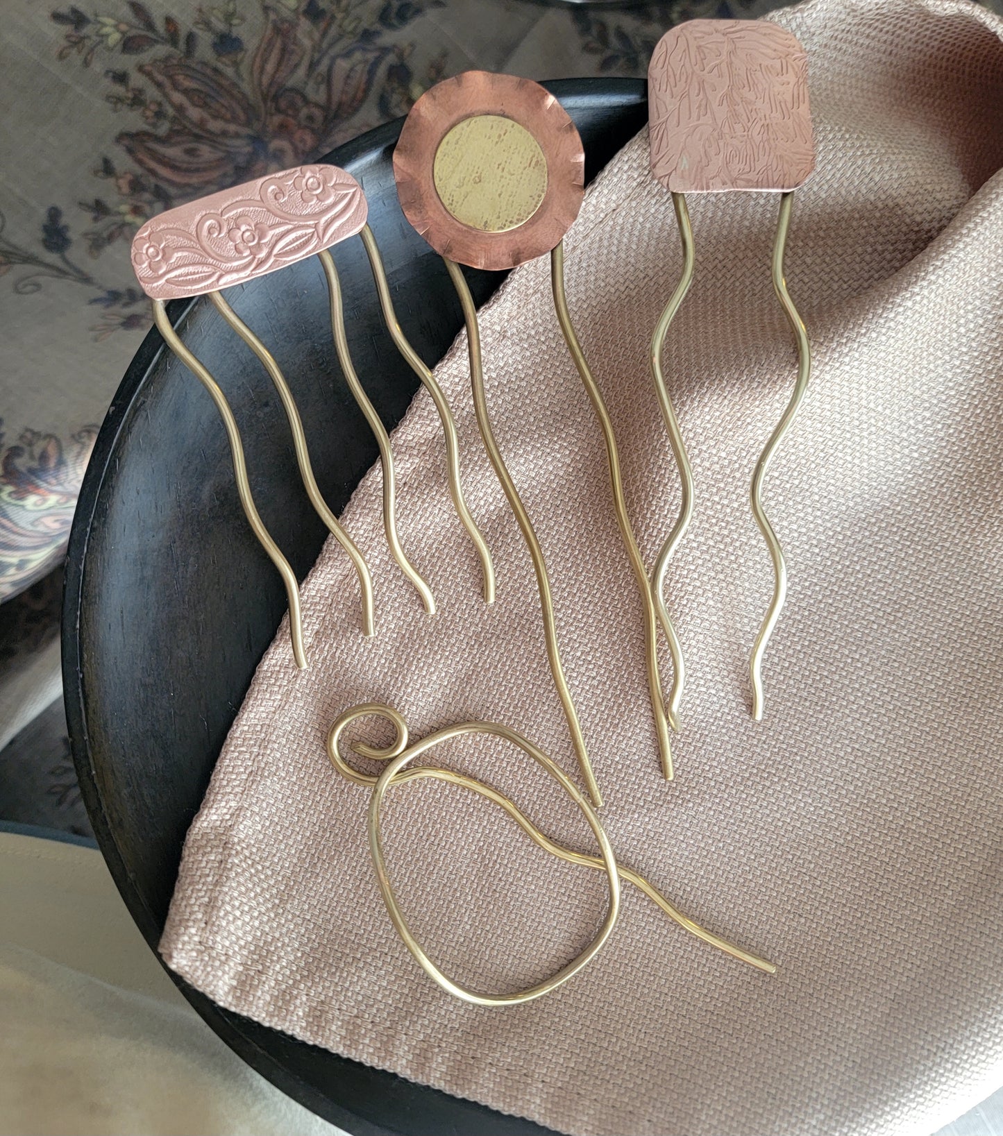 Three pairs of brass and copper hair pins and a bun holder on a wood plate with a beige cloth background